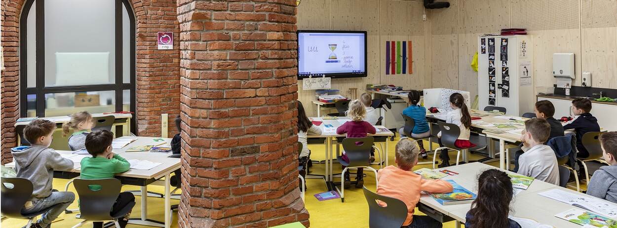 Kinderen kijken naar een digitaal schoolbord in klaslokaal met oude details van kerkgebouw nog zichtbaar.